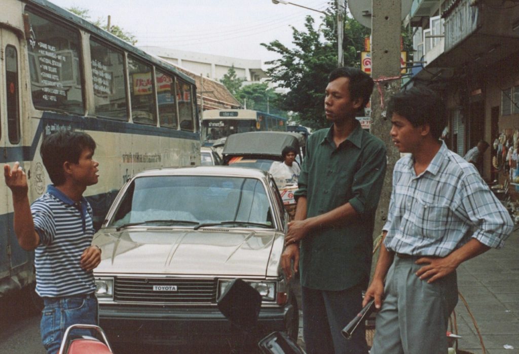 A photo of 3 young Thai con-men standing on a busy Bangkok street.